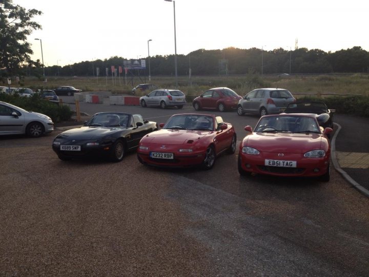 A bunch of cars are parked in a lot - Pistonheads - The image depicts a parking lot with four convertible cars parked back-to-back, each in a different color: red, black, silver, and pink. The cars are parked on a gravel surface, and in the background, there are various signs and a tall floodlight illuminating the area. The parking lot appears to be empty, except for these four cars, which suggests a quiet and relaxed atmosphere.