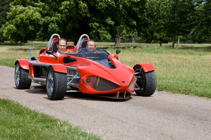 Pistonheads Manor Motors - The image captures a scene on a dirt road, where two men are seated in what appears to be a motorized kart or three-wheeled vehicle. The vehicle is characterized by its bright red color and an elongated body shape. It's parked on a gravel path, with the two men in long-sleeve white shirts attentively looking at something off-camera. The setting seems peaceful with a lush green background.