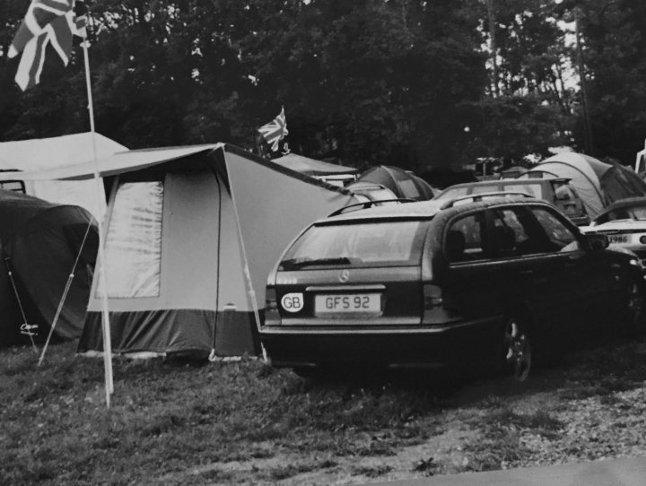 What private plates do you have? - Page 68 - General Gassing - PistonHeads UK - The image shows a camping site with several vehicles parked around. A black car is prominently parked under a tent, while other cars are parked nearby. In the background, there's a structure that resembles a small cabin or hut, possibly part of the camping facilities. To the left of the frame, there's a flagpole with a Union Jack atop, indicating a possible British influence or location. The overall atmosphere suggests an outdoor recreational area where people come to enjoy nature and the outdoors.