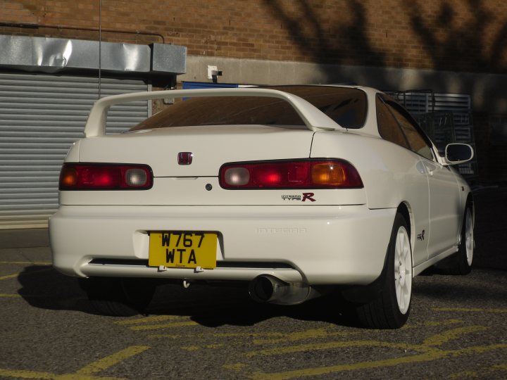 Pistonheads - The image captures a bright red sports car parked outdoors. The car's rear lights are visible, adding a vibrant splash of color against the car's white body. A black opaque door can be seen in the background, partially covered by a yellow curb. The license plate of the car reads "W76 WTA," indicating its registration. The car's sleek design is accentuated by the angle from which the photo is taken.