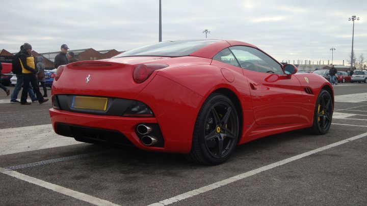 Service Oxford Sunday Pistonheads - The image depicts a vibrant red Ferrari car parked in a parking lot. The car's glossy finish reflects the surrounding environment, and the license plate is easily visible. Its two-door coupe design and yellow badge on the back are typical features of Ferrari sports cars. In the background, there are several other vehicles and a few blurred figures of people walking, giving a sense of scale and location, possibly at an outdoor event or near a popular destination.