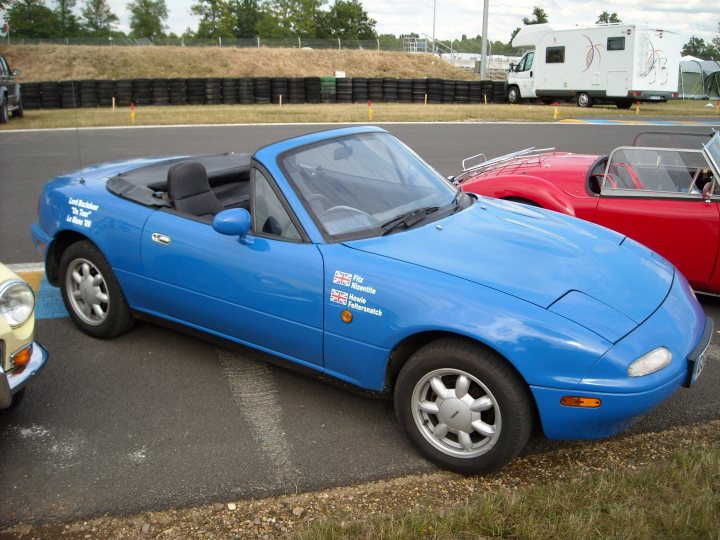 Pistonheads Stick Plates Traditional - The image presents a scene in a parking lot where a blue sports car, adorned with British and American flags, is parked. The car's sleek design is evident, with a convertible top, two doors, and alloy wheels. The backdrop features a track with a white camper van parked, along with several other larger vehicles and trailers. The car, with its distinctive colors and style, stands out prominently in this setting.