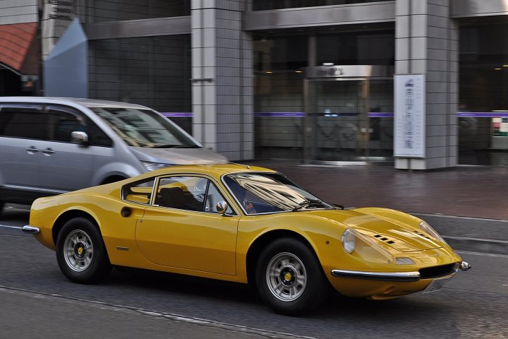 Supercars spotted, some rarities (vol 6) - Page 23 - General Gassing - PistonHeads - The image is a daytime street scene featuring a bright yellow sports car moving along a curved road. The car, a convertible with visible air intakes, appears to be the primary focus of the picture. It's hard to gauge the car's model or brand exactly, but based on the body language, it could be a Lotus or similar. To the right of the car, there is a signboard in another language, mounted on a grey wall which seems to be the facade of a building. In the foreground, a portion of a van is partially visible, and there are additional street elements like paving, clothing on a line, and a person's head on the street. The style of the image seems to be a real-life photograph.