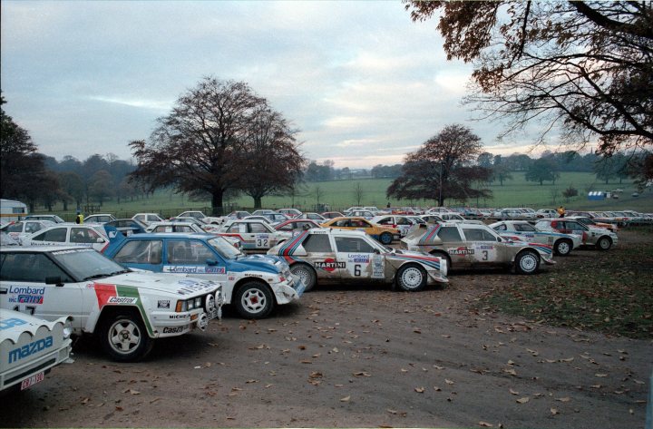 1985 RAC Rally Pics - Lancia Delta S4, 6R4 etc - Page 1 - General Motorsport - PistonHeads - The image shows a large lot filled with multiple white and colorful cars parked side by side. The vehicles are adorned with several stripes: red, white, and green. The cars appear to be rally cars or race cars, given the prominent stripes and the overall setting. The lot is large, with ample space for the vehicles, and it is lined with trees. The sky overhead is overcast, suggesting it might be chilly or cool outside. There is a sense of organized chaos, with the varied designs and the dense parking of the cars. Overall, the image gives a sense of anticipation, likely that of a race or driving event.