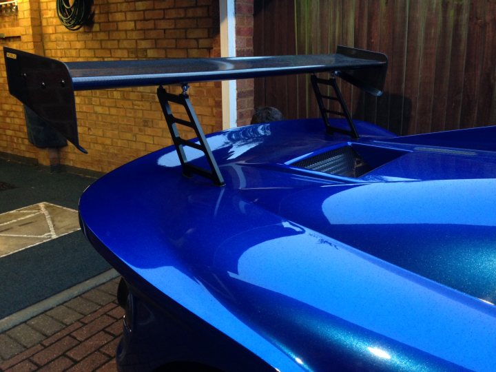 High Downforce Wing Risers - Page 1 - Noble - PistonHeads - The image showcases a blue sports car. The car's roof spoiler is striking, painted in a contrasting black color to match its wheels. A black grill on the hood adds to the car's aggressive appearance. The vehicle is parked on a brick driveway, with a red brick wall as a backdrop. The placement of the car in the frame suggests a focus on its design and color.