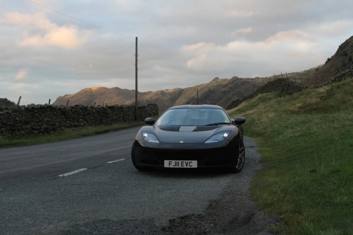 black evora spotted - Page 1 - Evora - PistonHeads - In the image, a black sports car is parked on the side of a road. The road is lined with wooden fence posts and flanked by grassy areas. In the distance, there are majestic mountains, adding a serene backdrop to the scene. The car's striking color contrasts with the natural tones of the roadside environment, making it the focal point of the photo. The photo captures a calm and peaceful moment, with the car inviting one to imagine a leisurely drive amidst the stunning scenery.