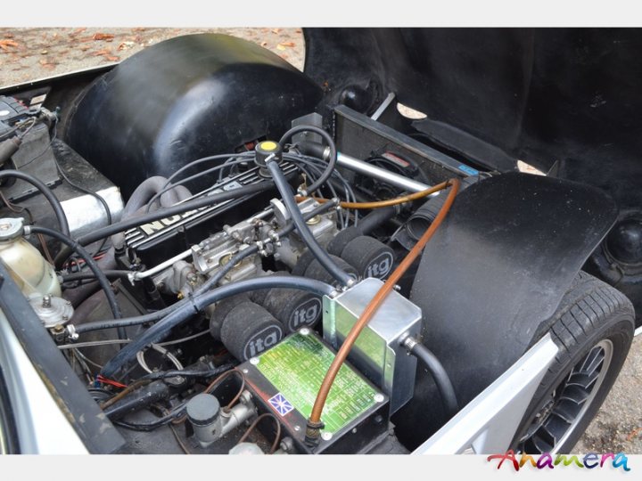 Early TVR Pictures - Page 36 - Classics - PistonHeads - The image shows a complex engine compartment of a vehicle, which appears to be an American car. The engine is open, and the various parts and wires are visible. The colors contrast with the predominantly dark engine, with the noticeable green and red components. The lens flare indicates sunlight, suggesting the photo was taken outdoors. The British flag sticker implies the car's likely origin or ownership.