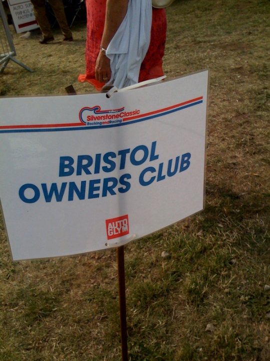 Grid Pistonheads Girlspit Babes - This image features a sign for the Bristol Owners Club. The sign is white with text that reads "BRISTOL OWNERS CLUB" in a combination of blue and red fonts. The sign also has logos for the Silverstone Classics and the Autoclassics, indicating the event venue, probably a racing event. The sign is placed on a grassy area, suggesting an outdoor setting, likely at a racing event or a classic car show. There's a metal pole in the foreground holding up the sign.