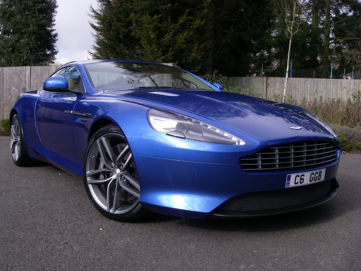 Can't wait till mid March!! - Page 2 - Aston Martin - PistonHeads - The image shows a sleek blue sports car parked on the side of the road. The car's body gleams under the sunlight, and it's positioned at a three-quarter angle, allowing a clear view of the wheels. The sky is overcast, suggesting a cool or cloudy day. There is a fence in the background, and some greenery, hinting at a suburban setting. The car's license plate is visible and reads "C6 GGB".