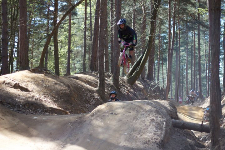 Picturesnot Pistonheads Bikes Kids - In the image, a young boy is actively engaged in mountain biking. He's skillfully navigating a trail across a dried-up dirt expanse. The boy is airborne, jumping over a little hill, his body leaning with the momentum of the jump. His bicycle is below him, momentarily suspended in midair. The setting is a scene of rugged nature, with trees scattered along the trail, providing a stark contrast to the boy's vibrant energy. The boy is in the center of the image, drawing the viewer's attention, while multiple other bikers can be seen in the background, adding depth to the scene.