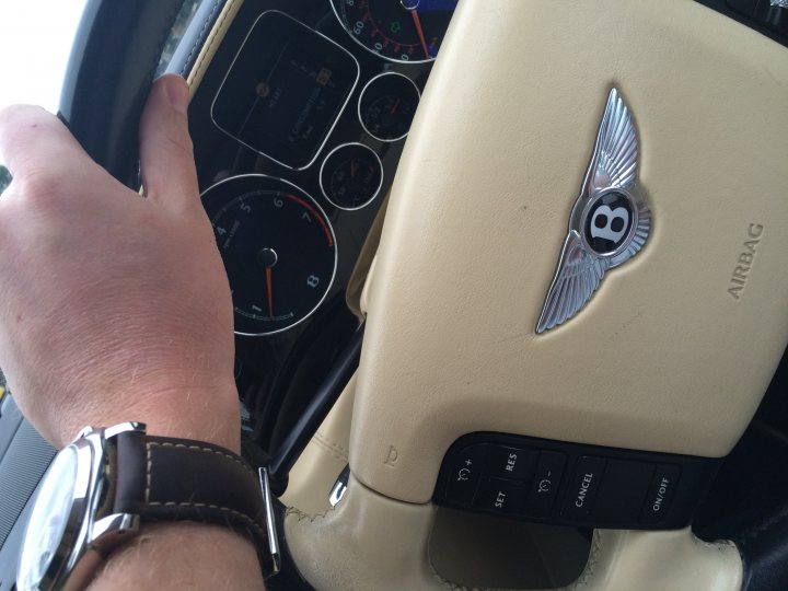 A person holding a cell phone in their hand - Pistonheads - The image shows a close-up view of the steering wheel of a luxury car, likely an Audi, as indicated by the dual-ring logo in the center. The steering wheel is prominently positioned in the foreground and occupies a large portion of the image. The car's dashboard can be seen in the background with various gauges and controls, including a clock and what appears to be a radio or multimedia system. There's a visible reflection in a shiny silver watch, which further becomes the focal point in the lower foreground, held by a hand across from the camera. The angle suggests the driver might have just brought their hand from holding the steering wheel back to normal driving position.