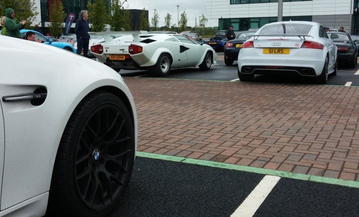 A white car parked next to a parking meter - Pistonheads - The image captures a scene in a parking lot filled with various types of cars. A white sports car is prominently parked, its sleek design contrasting with the surrounding cars. In the foreground, the front end of a luxury sedan is visible, its black tire representing the car's presence in the scene. A few blocks away, a red sports car adds a touch of color to the otherwise monochrome setting. A person is seen observing the scene, adding a human element to the otherwise mechanical landscape. The sky overhead is overcast, casting a soft light on the scene below.