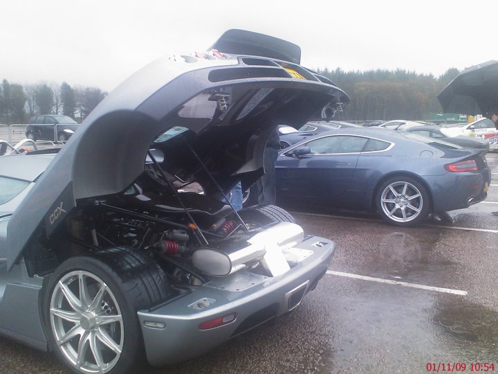 Pistonheads - The image presents a unique scene of a grey car with its trunk open. The car's hood is propped open, revealing the engine underneath. Rain slicked the surface of the area, reflecting the wetness in the surroundings. In the background, there is another car partially visible, and an umbrella can be seen at the edge of the frame. The overall scene suggests that some kind of work, possibly maintenance or inspection, is being carried out on the open-trunk car.