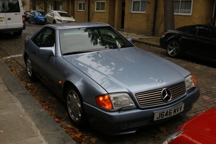 sl r129 anyone taken the risk on a 3/4k car - Page 20 - Mercedes - PistonHeads - The image features an urban scene with a silver Mercedes parked on the side of a brick road. The car is positioned parallel to the curb, but it appears to be parked too close to the center line, indicating a possible traffic violation. The road is lined with vehicles, highlighting the congestion typical of city streets. Buildings are visible in the background, suggesting that this may be a densely populated area. The presence of different types and styles of cars also indicates a multicultural urban environment.