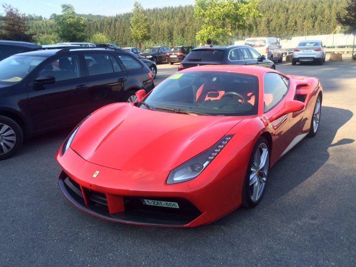 A red car is parked in a parking lot - Pistonheads - The image features a vibrant red Ferrari car parked on a paved lot. The lot is outdoors and filled with various vehicles, including sedans and other luxury cars. The Ferrari is positioned near a black hatchback, creating a contrast in color. It has black side mirrors and a matching hardtop convertible top, giving it a sleek and sporty appearance. The sunlight is casting a soft shadow to the left of the car, indicating it is late afternoon or early evening. The atmosphere suggests a warm, pleasant day.