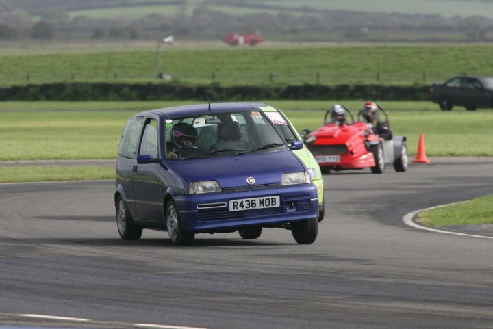 Pembrey Track September Day Pistonheads - The image captures a thrilling scene at a race track, featuring a small blue car and a powerful red car on a curvy track. The small blue car appears to be in the lead, expertly navigating the corner. The red car, equipped with number 43, is in close pursuit, demonstrating the intense competition of the race. There is a sense of speed and excitement in the air, emphasized by the pristine condition of the track and the professional appearance of the cars and their drivers. The background hints at a vast race track with ample open space, possibly near a rural area, adding to the overall spectacle of the race.