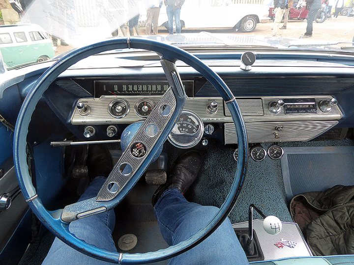 Pistonheads - The image showcases a vintage car interior, dominated by an old-fashioned steering wheel. The dashboard is adorned with various gauges and knobs, indicative of the car's era. A person can be seen sitting in the driver's seat, adding a human element to the scene. The car appears to be parked indoors, as suggested by the ceiling and artificial lighting. In the background, a glimpse of another vehicle is visible, hinting at a larger collection or exhibition.