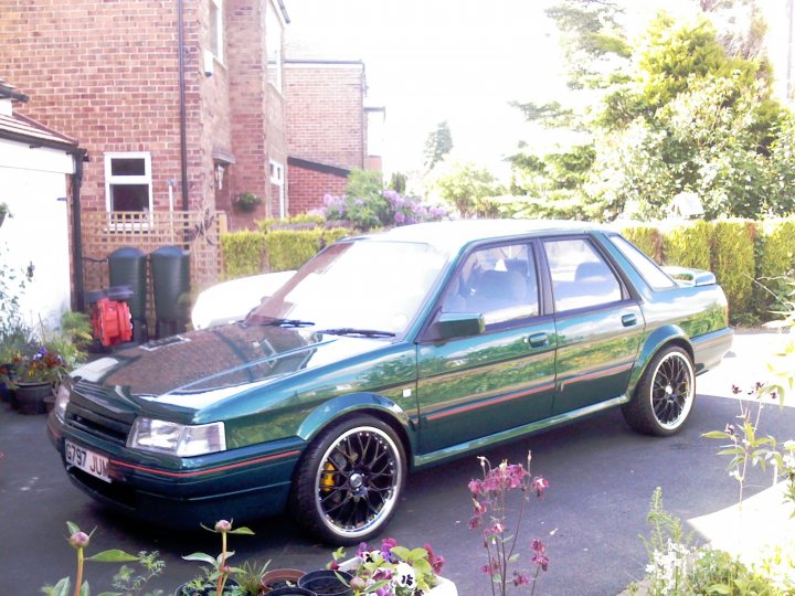 Turbo Montego Pistonheads - The image features a dark green-colored saloon parked in a driveway of a red brick house. The car is sleek and shiny, suggesting a well-maintained condition. The house itself has a white garage that matches the red brick exterior. On the right side of the driveway, there's a neatly trimmed hedge. The surroundings include other plants and decorative items, such as potted plants, creating a colorful and vibrant atmosphere. The image presents a harmonious blend of man-made structures and nature.