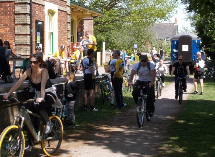 A group of people riding bikes down a street - Pistonheads - The image showcases a lively scene of a group of bicyclists, dressed in casual and sporty attire, congregated outside a building. They are lined up, with some people sitting on small chairs, while their bicycles are propped up on the path. Further back, the path seems to be a grassy area, and in the distance, a blue truck and some individuals can be spotted. The building they are at appears to be a brick structure with a large door. The overall atmosphere is vibrant and communal.