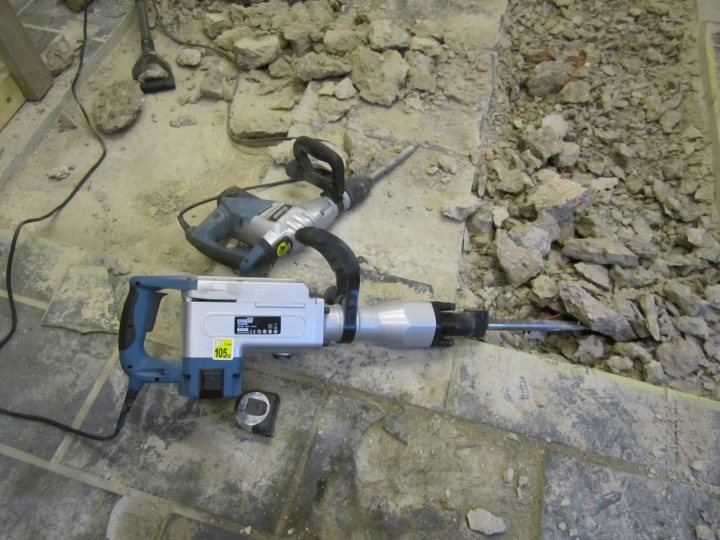 Pistonheads - The image displays a heavy-duty battery-operated rotary hammer power tool resting on a construction site concrete floor. The tool has a silver body and a blue grip, indicating it is from a particular brand, possibly Sigma. There are visible battery packs on the front, suggesting it's a cordless drill with quick-change attachments. Scattered debris and dust, along with a piece of string or metal, cover the ground, indicating ongoing construction work. The photo shows a dusty, industrial setting.