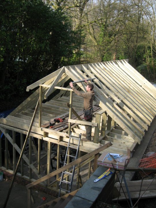 Finally the beginning of some decent petrol-head parking! - Page 1 - General Gassing - PistonHeads - The image features a construction site with a person working on a roof. The individual, who is wearing a dark shirt, is standing on a ladder, using a tool to secure the wooden shingles to the roof. The building structure is visible underneath, with visible support beams and wooden decking. The surrounding environment includes greenery, suggesting a quiet, possibly rural location.