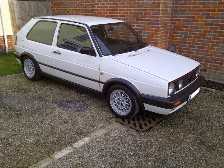 New toy :D - Page 1 - East Anglia - PistonHeads - The image displays a vintage white car parked on a driveway adjacent to a building with a brick facade. The vehicle has black trim around the body and black lower panels. The side windows are tinted, and there's a collar with a perimeter of foil on the lower side of the car, commonly used to prevent dents. The car features light-colored alloy wheels, and instead of a standard exhaust pipe, there is a custom-designed silver half-pipe near the rear wheel. There is a small branch leaning against the car. The scene might suggest a residential area, and the overall impression is that the car is well-maintained and possibly cherished as a classic or collector's item.