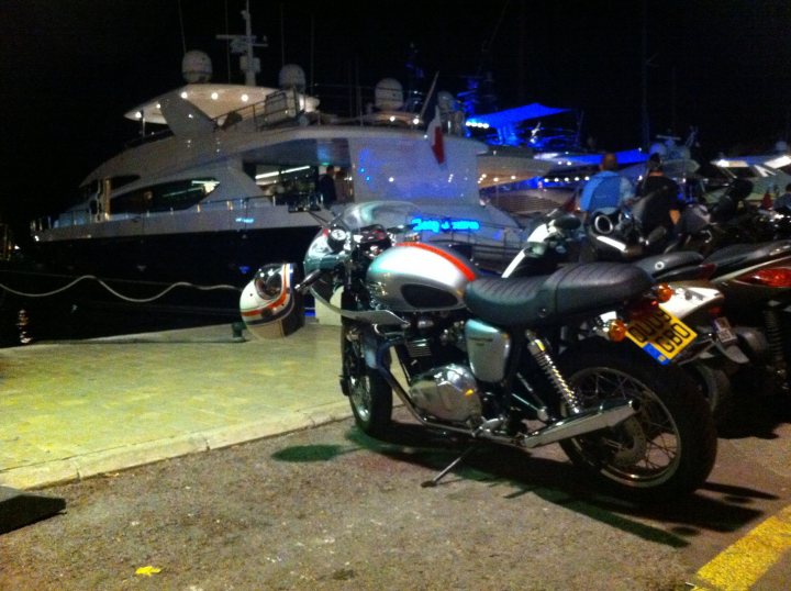 A row of motorcycles parked next to each other - Pistonheads - The image shows a parking area at night with boats visible in the background. The main subjects in the foreground are a parked motorcycle and a bicycle. The motorcycle has a prominent helmet, an orange and silver gas tank, and a leather seat. The bicycle is positioned between the motorcycle and the motorcycle owner's legs, suggesting the owner is standing nearby. The lighting in the image is primarily artificial, and the location appears to be a boat harbor due to the presence of boats in the backdrop.