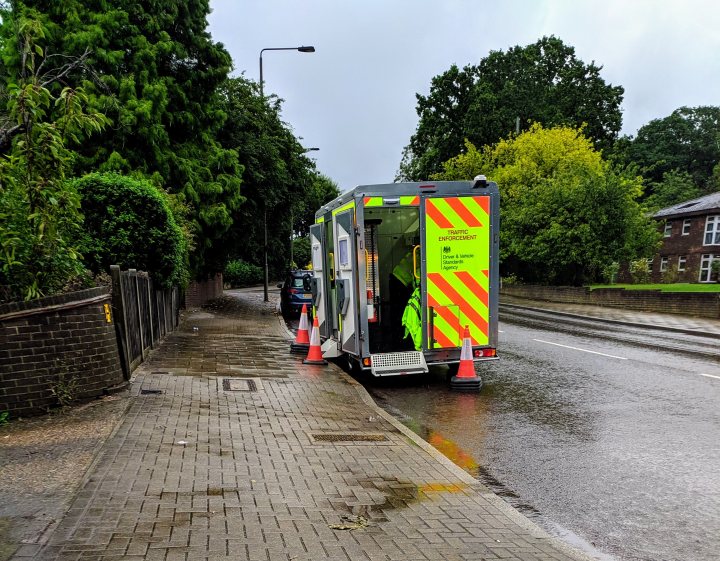 DVSA Spot Check - Page 1 - Speed, Plod & the Law - PistonHeads - The image depicts an outdoor scene on a rainy day. A large ambulance vehicle, marked with emergency service markings and reflective strips, is parked near the sidewalk. It has its doors open and appears to be stationary. The street is wet from recent rain, and there's a visible puddle next to the ambulance. A few cars are also present on the street, one of which is partially obscured by the vehicle. The sidewalk is bordered by trees and some buildings can be seen in the background.