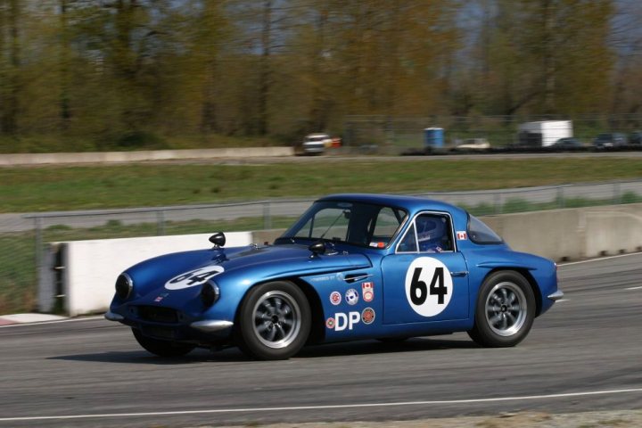 Early TVR Pictures - Page 67 - Classics - PistonHeads - This is a vivid image of a blue sports car, exterior number 64, racing on a track. The car is positioned in the lower right-hand corner of the frame, accelerating towards the right. The background features a lush green field, a partial track fence, and the back of a white trailer. The elevated angle of the shot captures the car in motion with the setting suggesting an outdoor motorsport event.