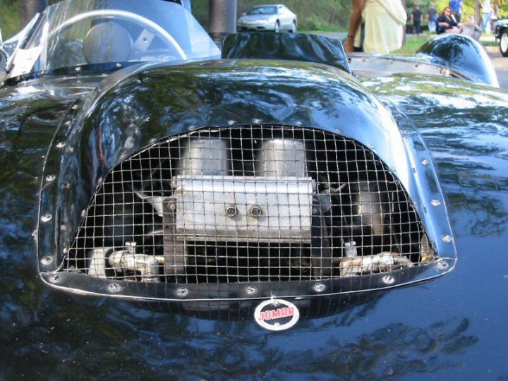 Early TVR Pictures - Page 2 - Classics - PistonHeads - The image shows a classic racecar parked outdoors. The front of the car features two large, cylindrical exhaust pipes on either side of the grill. The car's body is painted in a glossy black finish, which shines under the light, reflecting the texture of the car's material. Behind the racecar, you can see a blurred background indicating it might be on a track or a driveway. The car's grill and the mesh of the hood's gauges are clearly visible. The photo effectively captures the old-world elegance of the vintage vehicle.