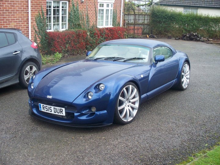 wheel arch flares like these.. - Page 1 - Cerbera - PistonHeads - The image showcases a parked blue sports car in the driveway of a house. The car has a sleek design with distinct headlights and a prominent front grille. It features a license plate with the text "RS16 DUR" and five-spoke silver rims. The house in the background has a brick facade and is partially obscured by a leafy tree.