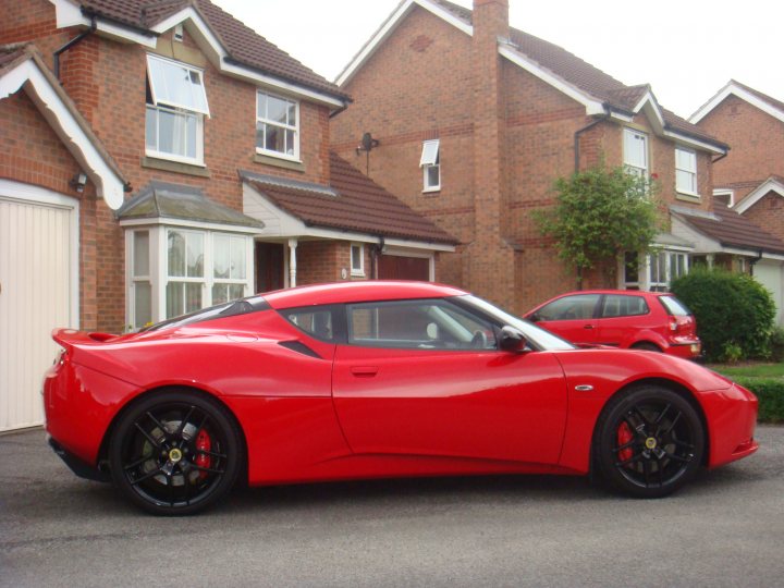 Finally some pictures!! - Page 1 - Evora - PistonHeads - The image showcases a vibrant red sports car parked on a residential street surrounded by brick houses with neatly maintained lawns and gardens. The car's sleek and aerodynamic design with its black rims and silver accents signify its powerful and sporty nature. The car is positioned directly under the buildings, and its shiny glossy finish reflects the light on the pavement, indicating that it is sitting still. The contrast between the vibrant red of the car and the more subdued brick houses and lush greenery create a striking visual.