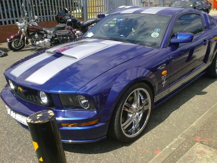 Atlantic July Sat Pistonheads Bbq Sports - The image shows a vibrant blue Ford Mustang car parked on the side of a road. It's a sunny day, and the car is casting a shadow on the ground, suggesting it's parked in the shade of a nearby structure. The Mustang is well-maintained, with a shiny exterior and shiny black rims. In the background, there are other vehicles and a fence. The car's sleek design and shiny appearance make it the focal point of the image.