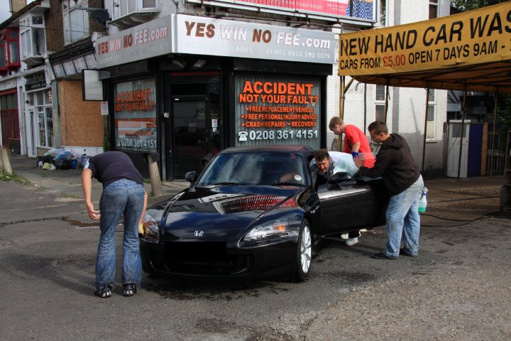 My S2000 review - Page 1 - Honda - PistonHeads - The image depicts a scene on an urban street. Three men are attending to a black car parked outside a business. The car has a clean and polished appearance, suggesting it might have just been washed. The car is positioned under a car wash sign, indicating that the washing service is being utilized. The three men vary in their tasks; one is maneuvering the car, another is bent over the hood, and the third is standing nearby, possibly overseeing the process. The car is located on a patch of wet ground, likely due to recent rainfall, which adds a glossy finish to both the car and the pavement.