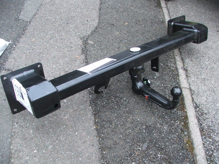 Pistonheads Bar Tow Fitting - The image shows a black car tow bar, consisting of a straight horizontal bar with a circular mirror on the left and a cylindrical connector on the right. The tow bar rests on what appears to be a paved area with road markings, indicating a road or parking lot. The bar itself is in the foreground and takes up most of the image, with a noticeable bolt pattern along one edge. There is no visible activity or movement in the image, and no vehicles are present except for the tow bar.