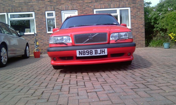 Pistonheads - The image is a photograph showing a red Volvo 740 sedan parked on a brick pavement in front of a house. The car has a distinctive front grille with the Volvo emblem and the headlights are visibly off. The license plate is visible and reads "N85 BJH." To the left of the car, there's another vehicle partially visible, and some off-road terrain is visible between the two. In the foreground, there's a green plant and a blue container near the bricks. The overall setting appears to be residential with a well-maintained exterior.