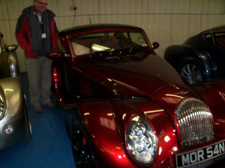 My new 4/4 lowline - Page 1 - Morgan - PistonHeads - The image features a man standing next to a classic red car. The man is dressed casually in a red and gray jacket. The car is sleek and well-maintained. Behind the man and the red car, there are several other vintage cars, indicating a setting where classic vehicles are displayed. Another person, smaller in the frame, is partially visible near the collection of classic cars.