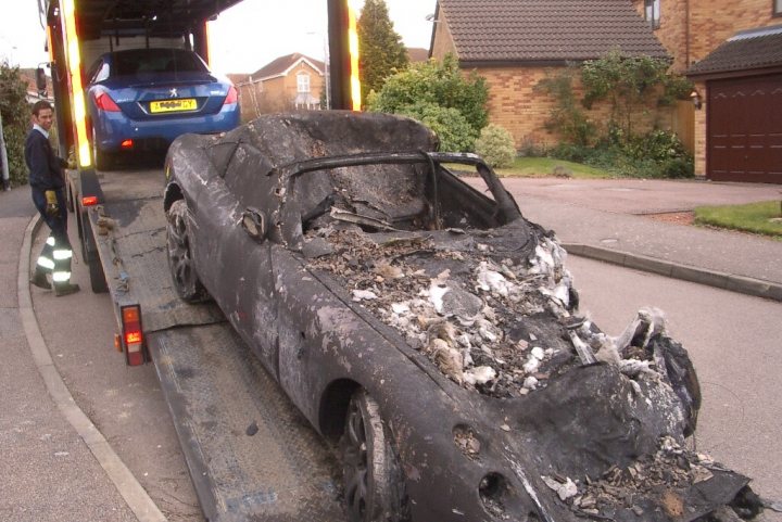 Dust Bites Pistonheads - The image shows a scene of a classic black car severely damaged with signs of fire and being crushed by a car carrier. The car is resting on the bed of the tow truck, and the surrounding area includes a brick house and a blue car in the background. The sun is shining, and the man in a striped jumpsuit is attentively observing the situation from the left side of the image. This appears to be a residential area with trees and grass visible alongside the road where the incident occurred.