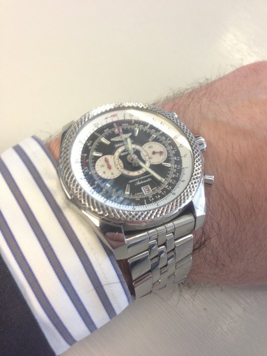 Let's see your Breitling.  - Page 10 - Watches - PistonHeads - The image features a silver wristwatch with a black bezel. The watch face displays a combination of black, silver, and white colors with various markings. The watch strap is partially visible with a pattern of silver and blue stripes. A blurred human hand and forearm are in the background, suggesting the watch is being worn. The image is close up, focusing on the watch, and the person's identity or arm color are not emphasized.