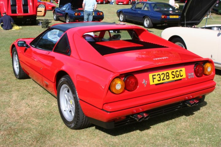 F353 TRY - Anyone know it? (328 GTS) - Page 1 - Ferrari V8 - PistonHeads - The image showcases an outdoor event, possibly a classic car show, where a vibrant red sports car stands out. This car is positioned prominently in the middle of the frame, drawing attention with its shiny exterior and shiny gold covers. The car has a unique shape, suggesting it's a convertible model, emphasized by a red top. Other cars can be seen in the background, adding to the atmosphere of a car show. The greenery and road nearby suggest the event is taking place in an open space or near a roadside.