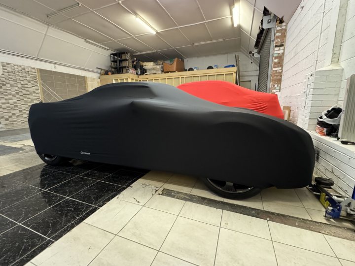 Pistonheads - The image depicts a car with a black tarp covering it, parked inside what appears to be an enclosed parking space or garage. The vehicle is positioned parallel to the back wall of the space, and its front end faces towards us, the viewer.