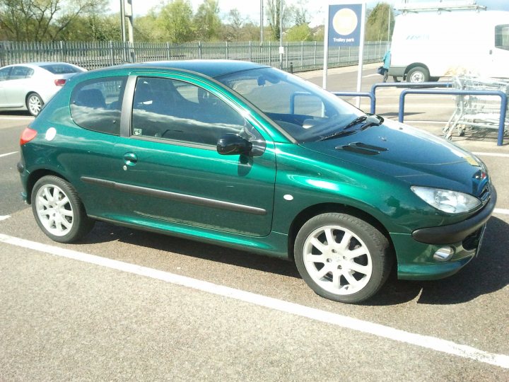 206 gti - Page 1 - French Bred - PistonHeads - The image is a color photograph depicting a green car in a parking lot. The vehicle is a compact, four-door car with the driver's side front door open. It features alloy wheels and a visible license plate at the front. The windows of the car have a slight reflection on them, and the exterior appearance of the car suggests that it is relatively new or well-maintained. In the background, there are other cars parked and a signboard with what appears to be a "Trolley Park" graphic, indicating the location might be a public parking area.