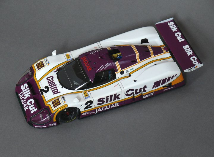 Tamiya 1/20th Jaguar XJR9-LM - Page 14 - Scale Models - PistonHeads - The image depicts a meticulously crafted model race car. It is primarily a white and purple, with the driver's side panel in a maroon and orange color scheme, suggesting the livery of the JaGa Racing team. The model contains intricate details such as brake calipers, wing end plates, and numerous sponsorship decals. Text running along the side of the car reads "SILK SLO CUT" with the JaGa Racing logo placement and "SILK GIR" on the rear wing end plate. The model sits against a muted grey background, emphasizing the vibrant colors and fine craftsmanship of the car model.