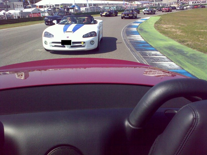 Pistonheads Jim Hockenheimring Clark - This image captures a dynamic scene at a race track. There are two white sports cars, one with a blue racing stripe, racing on the track. The perspective is from the passenger seat of a vehicle at the side of the track, with the car's windshield in the foreground. The track itself curves gently, reflecting the cars as they race. The setting suggests a professional racing event.