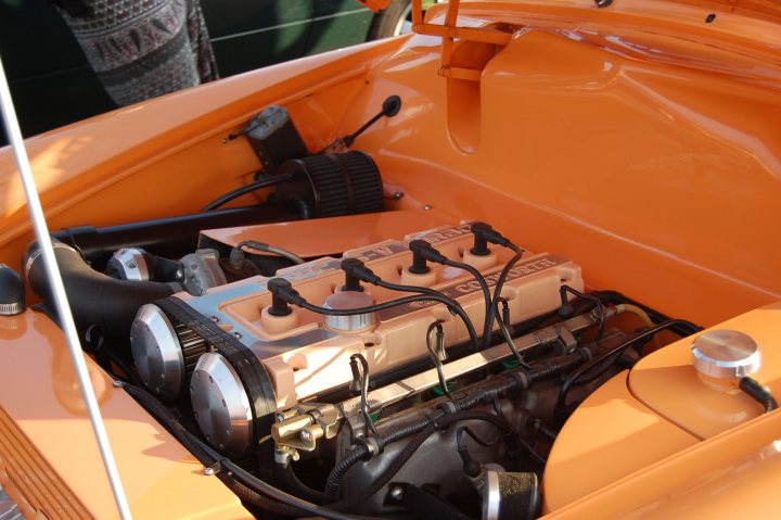 earls barton clasic car meet tomorrow april 24th  - Page 1 - Northamptonshire - PistonHeads - The image displays an open bonnet revealing the engine of an orange vehicle. The engine is complex, with a visible carburetor, and is surrounded by a web of black and white cables and wires. To the left, there's an exhaust pipe and a garage-like background is visible in the top left corner. The photo captures the intricate details of the engine, with accessories like a chrome shield under the hood. The vehicle appears to be a vintage or classic car based on the design and engine configurations typical of the 1950s or 1960s.