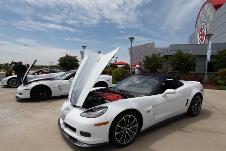 Corvette challenge what would you change? - Page 1 - Corvettes - PistonHeads - The image shows a white Corvette parked in the foreground, with its hood propped open revealing the engine. The Corvette is parked outdoors on a paved surface, likely at a car show or a similar event. In the background, there are several other individuals and a few cars parked nearby, contributing to a scene of a large gathering of car enthusiasts. The posters on the wall suggest a commercial setting, possibly a car dealership or a similar venue. The participants appear to be examining the cars or possibly socializing.