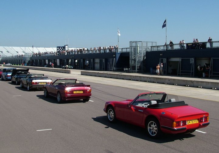 Pistonheads - The image shows a row of classic sports cars on a track, with people standing on a balcony or pit lane deck overlooking the scene. The cars are various colors, including red, black, and blue, and are positioned closely together as if ready for a race or event. The balustrade provides a clear separation between the viewing area and the track. The sky is partly cloudy, and the setting sun casts long shadows on the track.