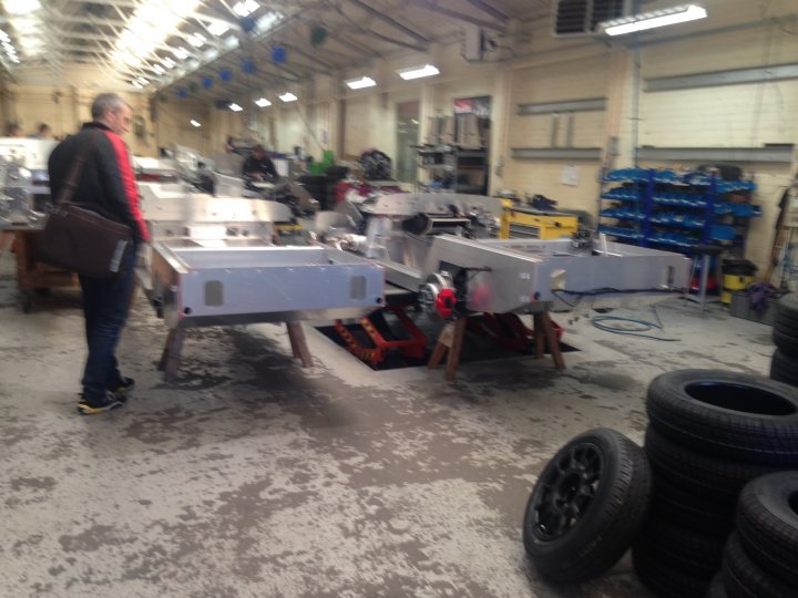 RE: Morgan Sunday Service 17/4/2016 - Page 5 - Sunday Service - PistonHeads - This image captures a scene from a garage or workshop. There are two large, metallic contraptions or machines on pedestals, one of which a man with a colorful jacket and a bag attached to his shoulder is standing near. The machines appear to be of a similar industrial type, suggesting that they might be related to a specific process or function. The background reveals an environment with various objects and possibly other machinery, reinforcing the setting as a workspace or production area.