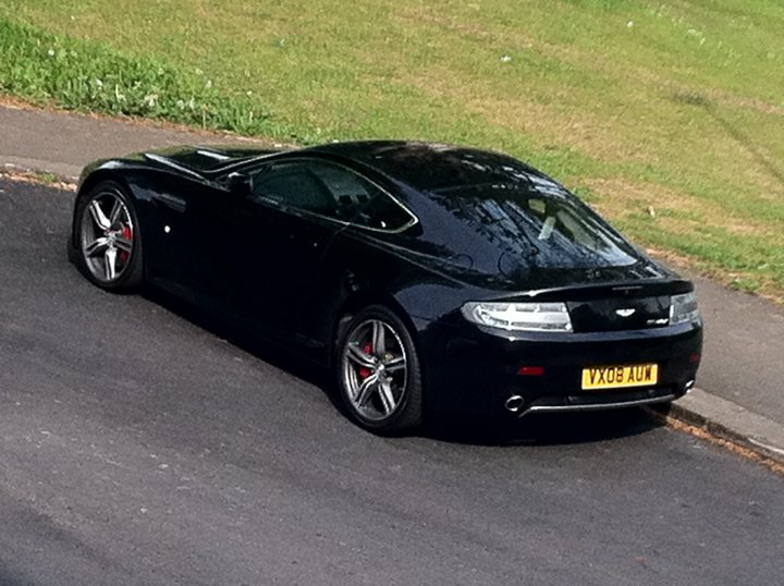 Bought Woo Hoon Pistonheads - This image features a black sports car parked on the side of a street. The car is positioned at a slight angle to the camera, showcasing its design and features. The vehicle has a sleek, low-profile design, indicative of the sports car class. On the front bumper, there is a yellow license plate displaying the code 'VX08 AGU'. The surrounding environment includes a sidewalk and what appears to be a grassy area, suggesting a suburban setting.