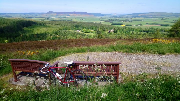 The "Photos From Today's Ride" thread. (Vol. 2) - Page 24 - Pedal Powered - PistonHeads UK - The image depicts a serene countryside scene. Dominating the foreground is a wooden bench, inviting and unoccupied, positioned slightly to the right of the center. To the left of this bench sits a solitary bicycle, parked securely. The background unfurls with rolling hills that stretch out into the distance under a clear blue sky. A white building nestles amongst these hills, adding an element of human presence to this pastoral landscape.